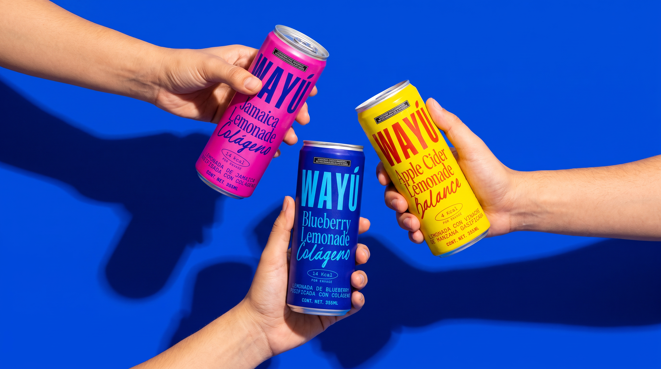 Manos brindando con latas Wayú Jamaica, Blueberry y Apple Cider Lemonade sobre fondo azul: misión Wayú